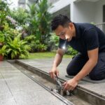 Vector control technician installing a stainless steel rat exclusion valve in a ground-floor drain during inter-monsoon season in Singapore.